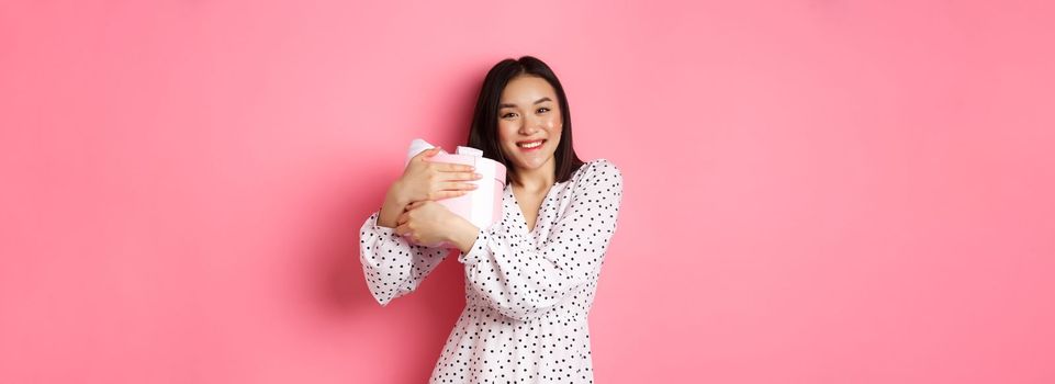 Lovely asian woman hugging her gift and smiling thankful, receive valentines day present, standing over pink background.