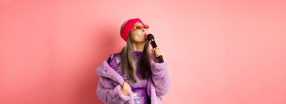 Stylish asian senior woman singing song, performing karaoke with microphone, standing in party outfit and faux fur coat against pink background.