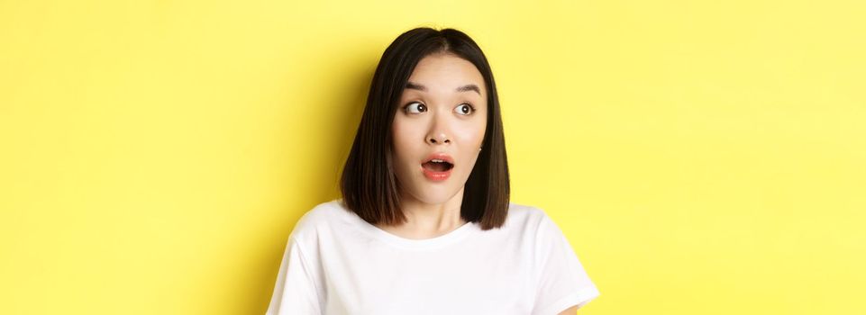 Close up of surprised asian girl drop jaw, gasping and looking left at logo, standing over yellow background.