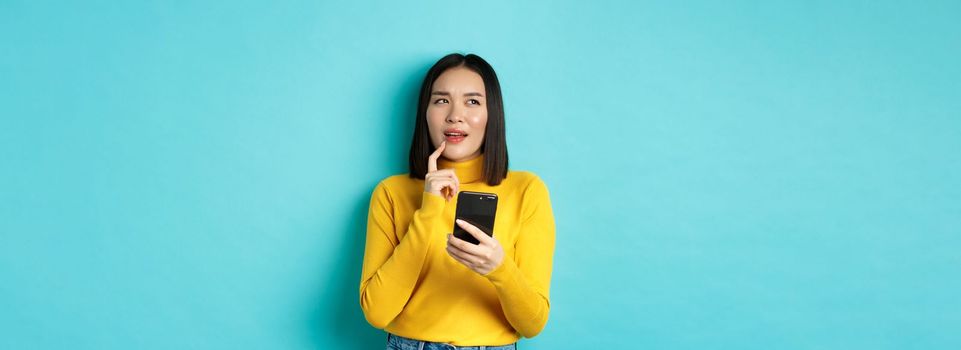 Pensive asian girl holding smartphone and thinking what to order online, standing over blue background.
