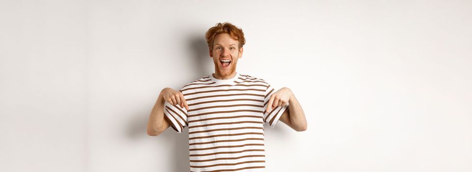 Enthusiastic young man with ginger hair and bristle showing advertisement, pointing fingers down and smiling, white background.