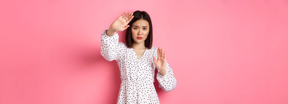 Timid and scared asian woman defending herself, raising arms in protection, victim being attacked, standing over pink background.