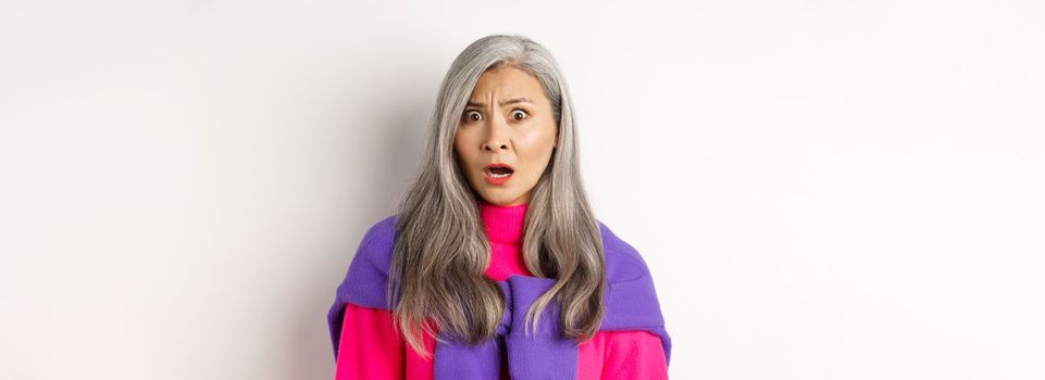 Close-up of shocked asian senior woman gasping, staring with confused face, looking with cringe, standing against white background.