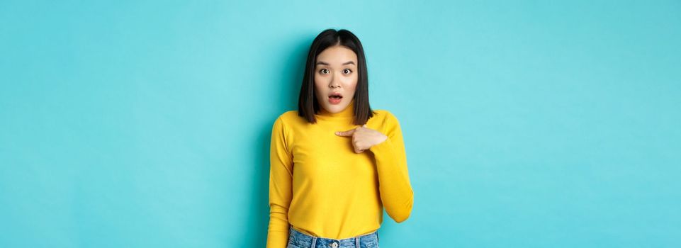 Image of surprised asian girl gasping and pointing at herself with disbelief, standing over blue background.