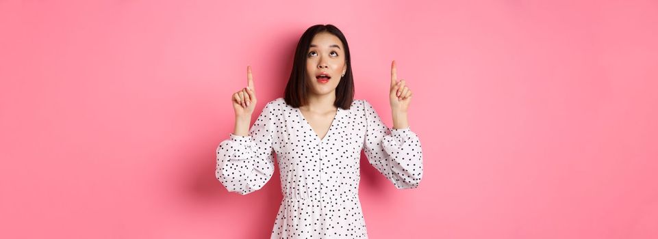 Image of beautiful asian girl in dress looking, pointing fingers up, gazing dreamy at promo offer, standing over pink background.