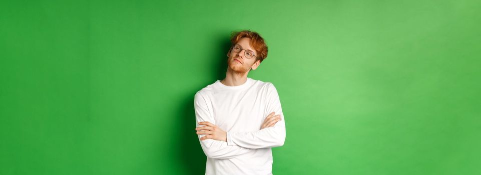 Confident young man with red hair, wearing glasses, looking smug at right side copy space, cross arms on chest self-assured, standing over green background.