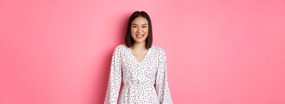 Happy korean woman in dress looking at camera, smiling and laughing with sincere expression, standing over pink background. Copy space