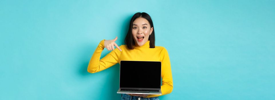 E-commerce and online shopping concept. Excited asian woman making presentation, pointing finger and showing blank laptop screen, standing over blue background.