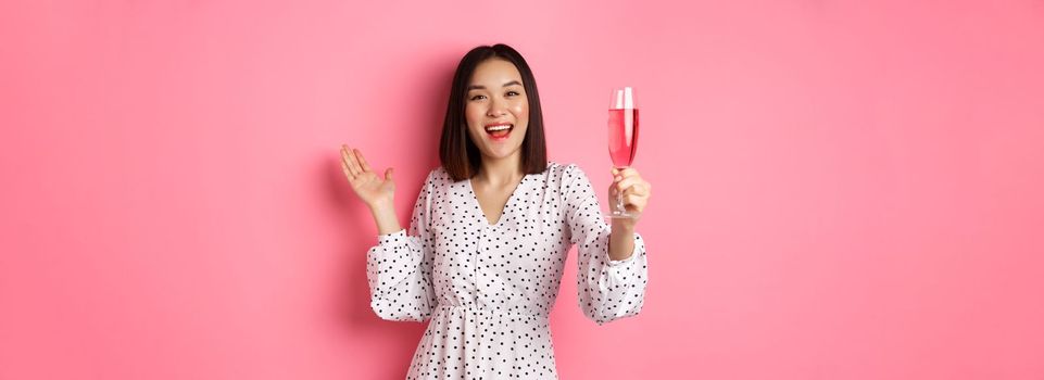 Happy asian woman celebrating, saying toast on party, raising glass of champagne and smiling, standing in dress over pink background.