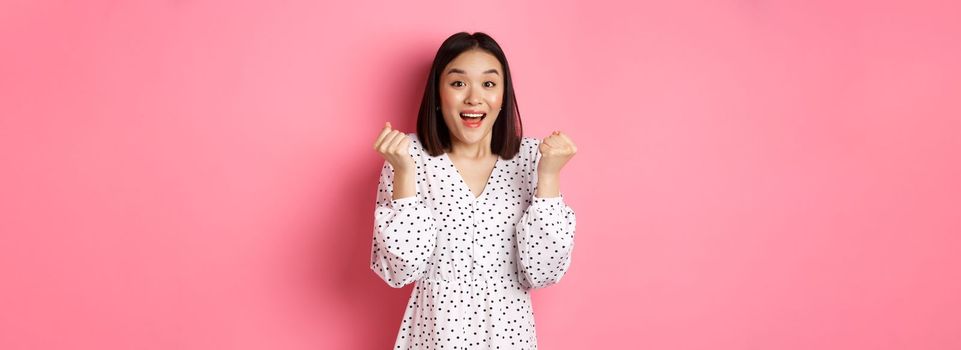 Image of beautiful asian woman winning, clench fists and saying yes with happy face, triumphing and celebrating victory, standing over pink background.
