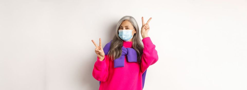 Covid, pandemic and social distancing concept. Cheerful and stylish asian senior woman wearing medical mask and showing peace signs, standing over white background.