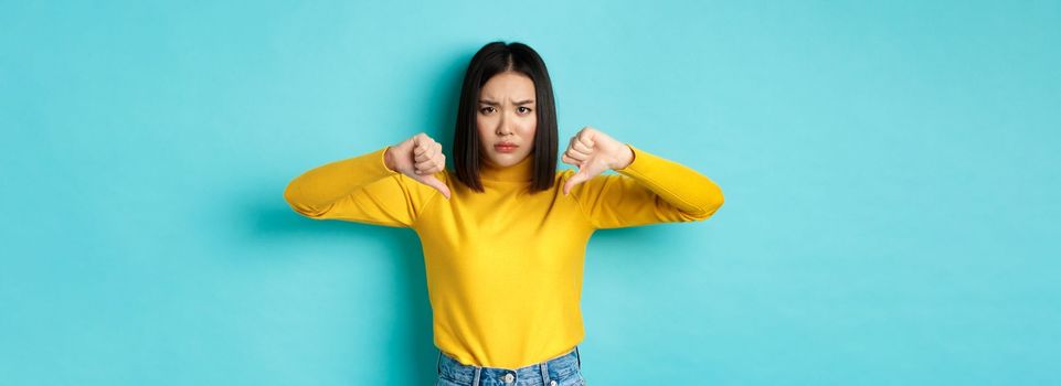 Disappointed asian woman frowning upset, showing thumbs down in dislike and disapproval, standing over blue background, staring at camera.