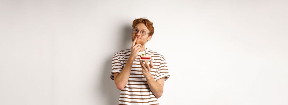 Holidays and celebration concept. Happy young man with red hair and glasses having birthday, holding cake with candle and thinking of b-day wish, standing over white background.