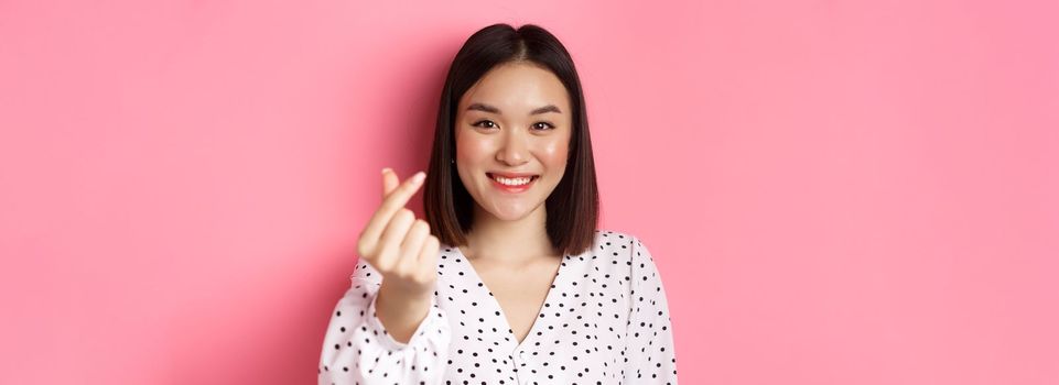 Beauty and lifestyle concept. Close-up of lovely asian woman showing heart sign, smiling and feeling romantic on valentines day, standing over pink background.