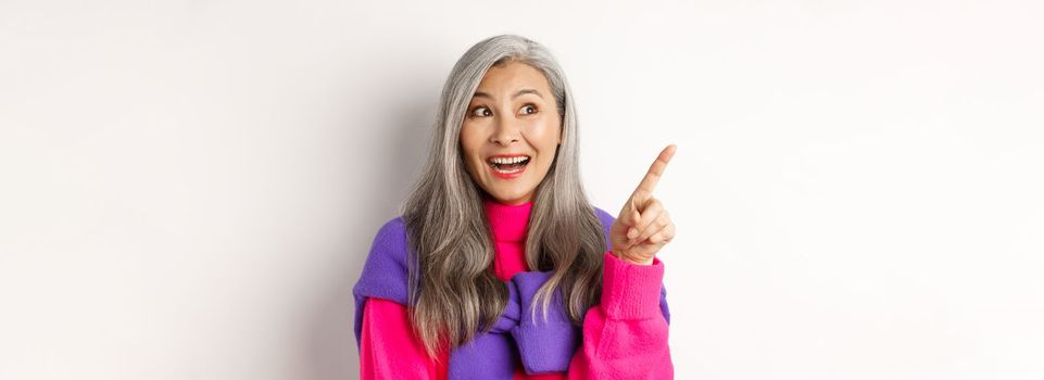 Close-up of stylish korean senior woman with grey hair looking and pointing upper left corner, showing promotion offer, standing over white background.