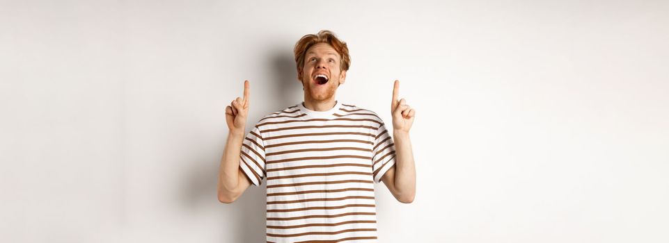 Fascinated redhead man looking with amazement and happiness, pointing fingers up, checking out something cool, standing over white background.