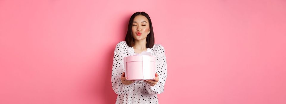 Cute asian girlfriend congratulate with valentines day, giving cute romantic gift in box, pucker lips for kiss, standing over pink background.