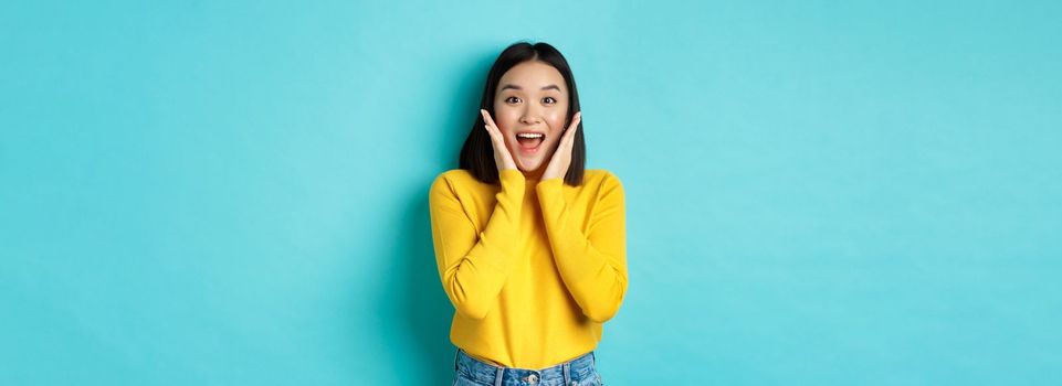 Portrait of pretty korean girl receive surprising news, looking amazed and happy at camera, standing over blue background.