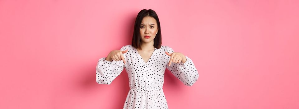 Disappointed asian female model pointing fingers down at promo offer, frowning and staring at camera upset, standing over pink background.