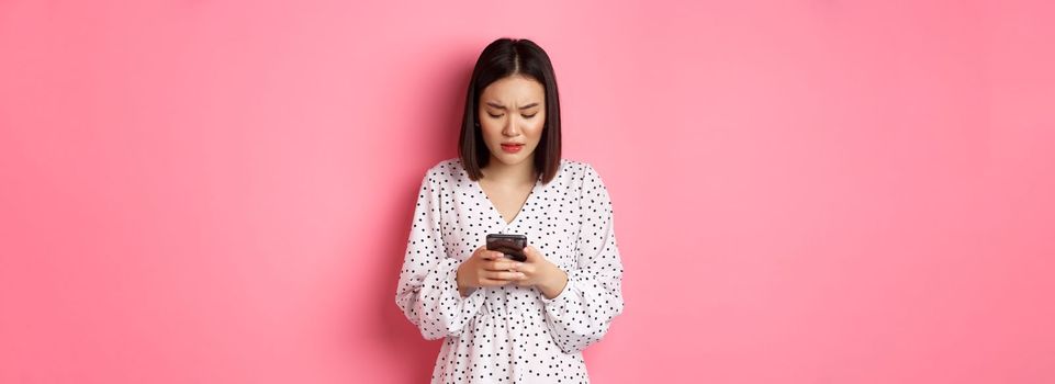 Nervous and concerned asian woman reading message on smartphone, looking worried, standing in dress over pink background.