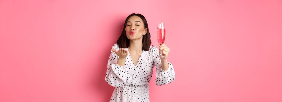 Romantic asian woman raising glass of champagne and sending air kiss at camera, celebrating and having fun, standing over pink background.