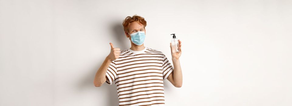 Covid-19, health and lifestyle concept. Smiling male model with red hair, wearing face mask, showing hand sanitizer and thumb-up, recommend to use antiseptic, white background.