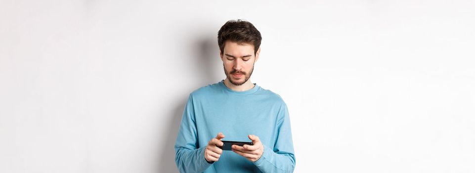 Male model playing video game on smartphone, looking serious at mobile screen, standing in sweatshirt over white background.