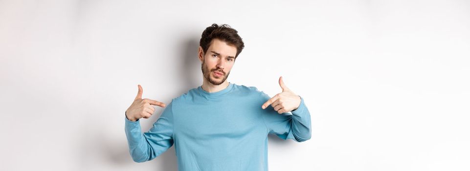 Young man with beard feeling confident, pointing at himself and making smug face, standing on white background cool and sassy.