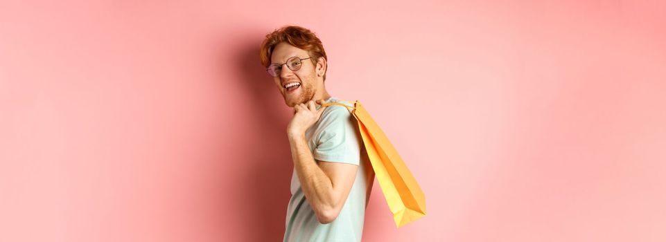 Carefree young man with red hair and glasses, walking with shopping bag over his shoulder and smiling, shopper buying presents, standing satisfied over pink background.