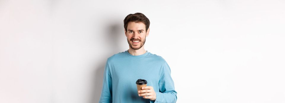 Cheerful man drinking coffee from cafe take-away and smiling, standing with paper cup in white background.