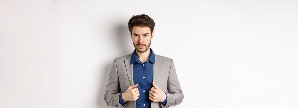 Confident young man with beard fixing suit and looking sassy at camera, feeling ready and determined, standing on white background.