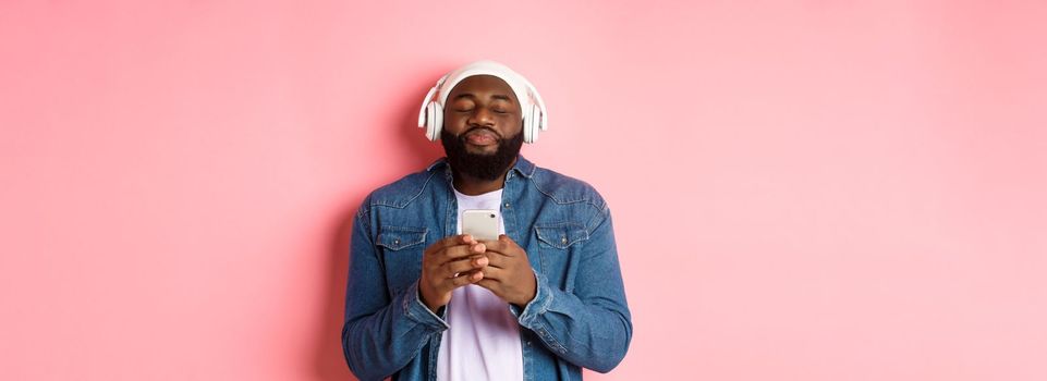 Delighted Black man enjoying awesome music, listening songs in headphones and holding smartphone, looking ecstatic, standing over pink background.