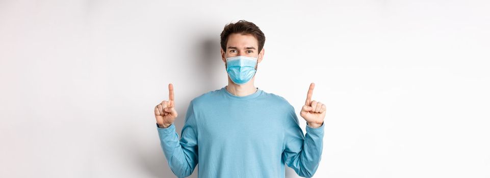 Coronavirus, health and quarantine concept. Handsome caucasian male model showing banner on top, pointing fingers up and wear medical mask, white background.
