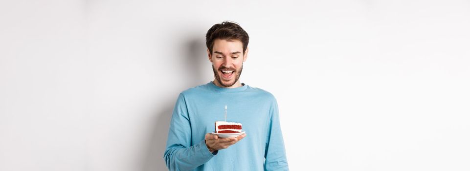 Cheerful man looking happy at birthday cake, celebrating bday, standing over white background.