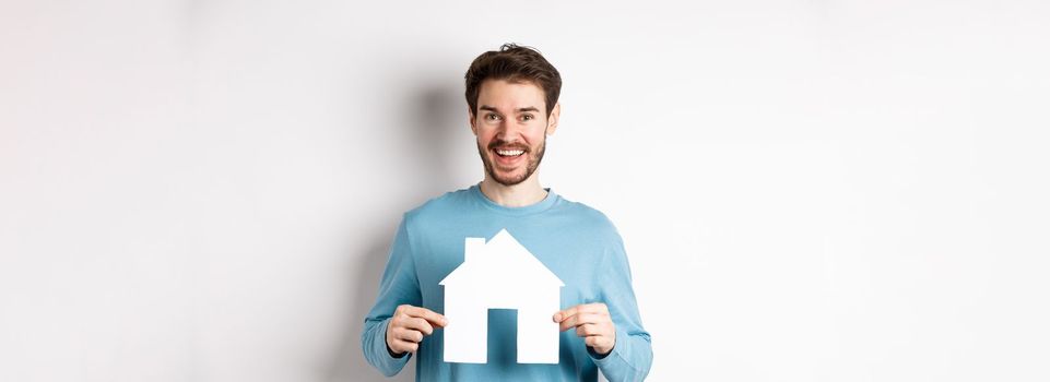 Real estate and insurance concept. Handsome modern man buying property, smiling and showing paper house cutout, standing over white background.