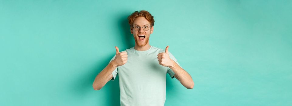 Excited young man with red hair, wearing glasses, showing thumbs-up and agree or praise something, smiling amazed and saying yes, standing over turquoise background.