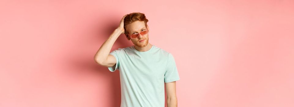 Handsome young redhead man in sunglasses, brushing hair with hand and looking smug and confident at camera, standing over pink background.