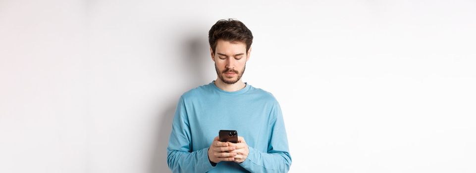 Serious young man reading message on smartphone, looking at mobile screen, standing over white background.