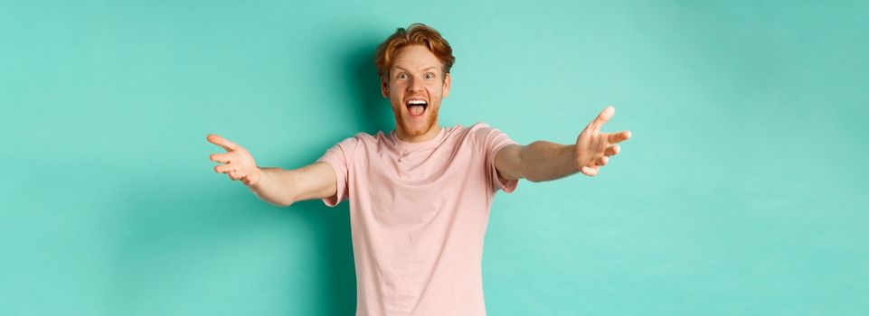 Friendly and happy young man with ginger hair, stretch out hands in warm welcome, reaching for hug and smiling joyfully, standing in t-shirt over mint background.