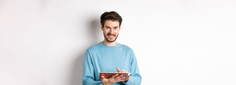 E-commerce. Smiling caucasian man using digital tablet and looking at camera, standing on white background.