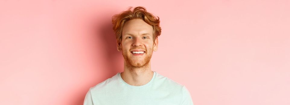 Happy young man with beard and messsy red haircut, smiling with white teeth and cheerful expression, standing over pink background.