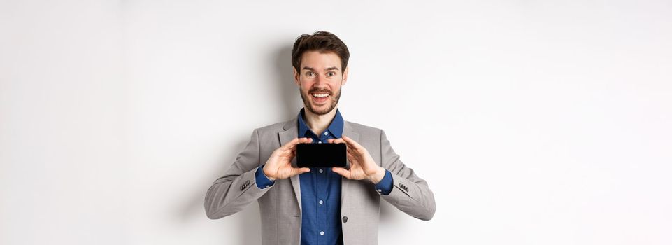 E-commerce and online shopping concept. Excited young businessman in suit showing empty smartphone, screen demonstrate app achievement, white background.