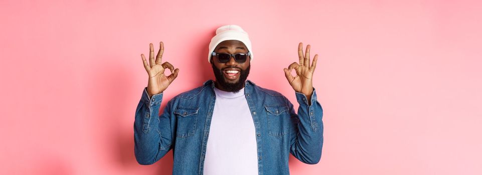 Image of cool african-american hipster man showing okay signs in approval, like something awesome, wearing sunglasses and beanie, pink background.