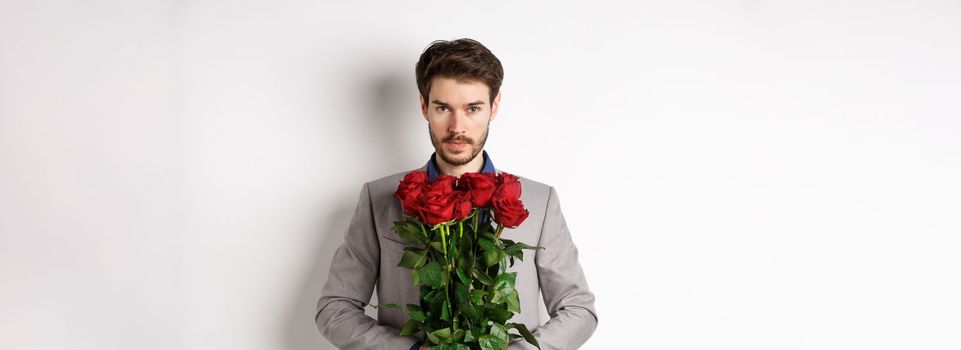 Valentines day and love concept. Confident and handsome man in suit holding bouquet of roses, looking at camera, standing over white background.