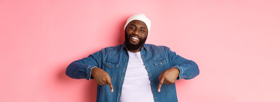 Happy Black man with beard, wearing hipster clothes, pointing fingers down and smiling, showing promo offer copy space, pink background.