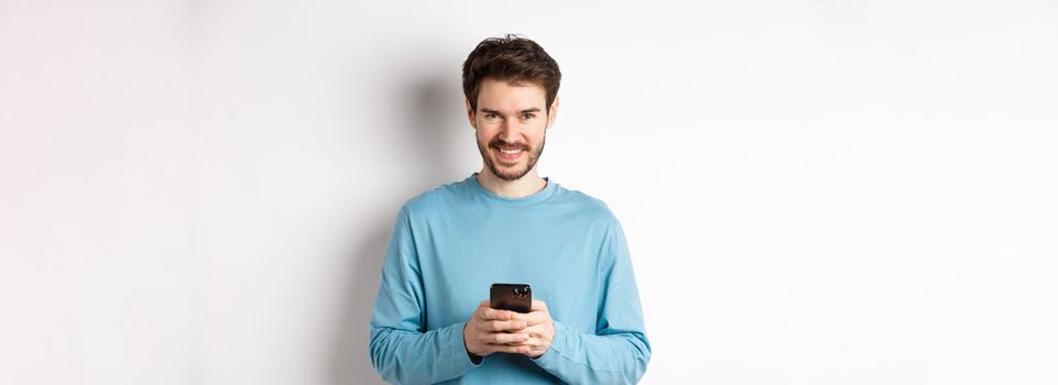 Smiling modern man using smartphone and looking pleased at camera. Guy in sweatshirt with mobile phone on white background.