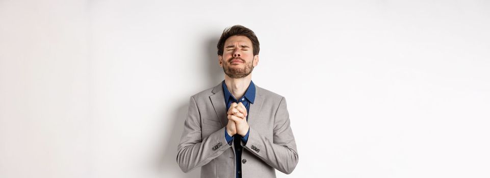 Nervous male entrepreneur in suit begging god, holding hands in pray with pleading expression, close eyes and make wish, asking for help, standing on white background.