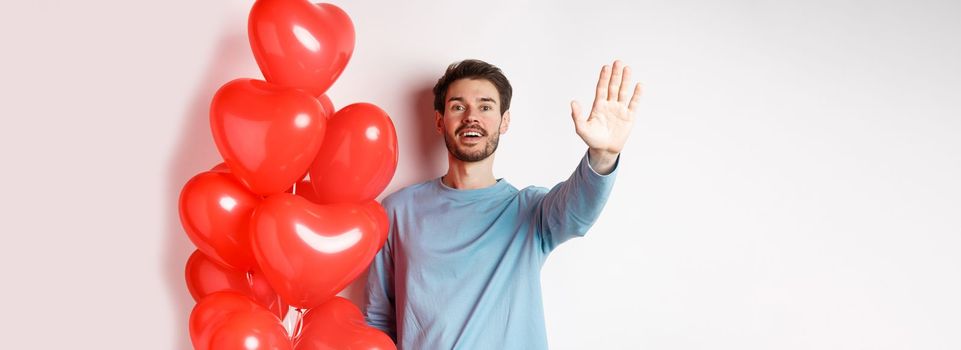 Handsome young caucasian man standing with romantic heart balloon and waving hand at lover, waiting for his date on Valentines day with cute surprise, standing over white background.