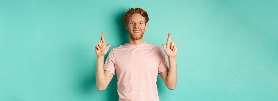 Optimistic guy with red hair and beard smiling, cross fingers for good luck and looking hopeful at camera, making a wish, standing over mint background.