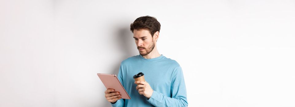 Man reading news online in digital tablet, looking serious at screen while drinking coffee from paper cup, standing over white background.